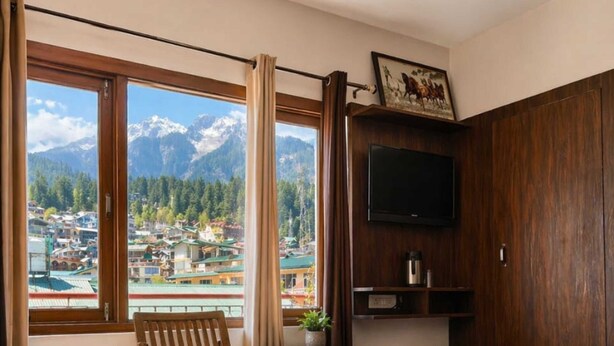 A bedroom with a bed, a TV, a wooden chair, a window with a mountain view, and a small table with a plant.