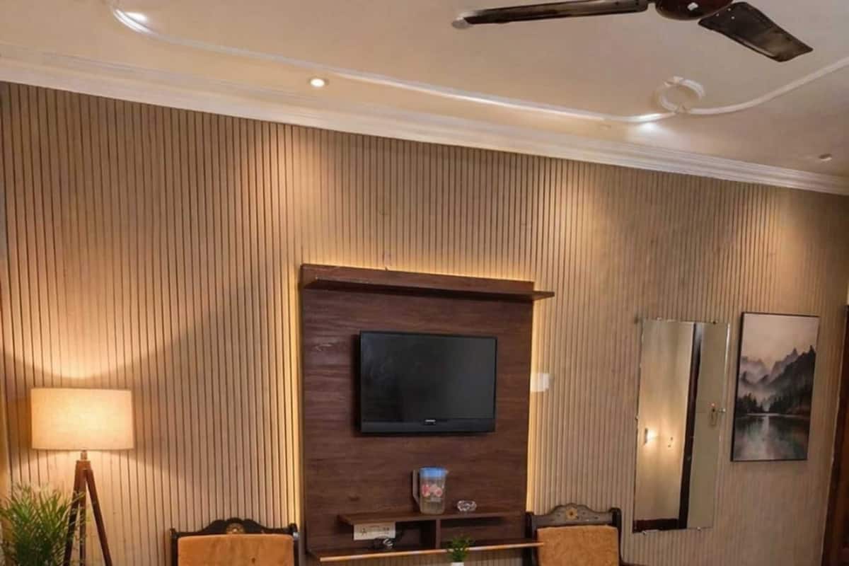 A hotel room with a bed, two armchairs, a small table, a TV mounted on a wooden panel wall, a ceiling fan, and a potted plant.