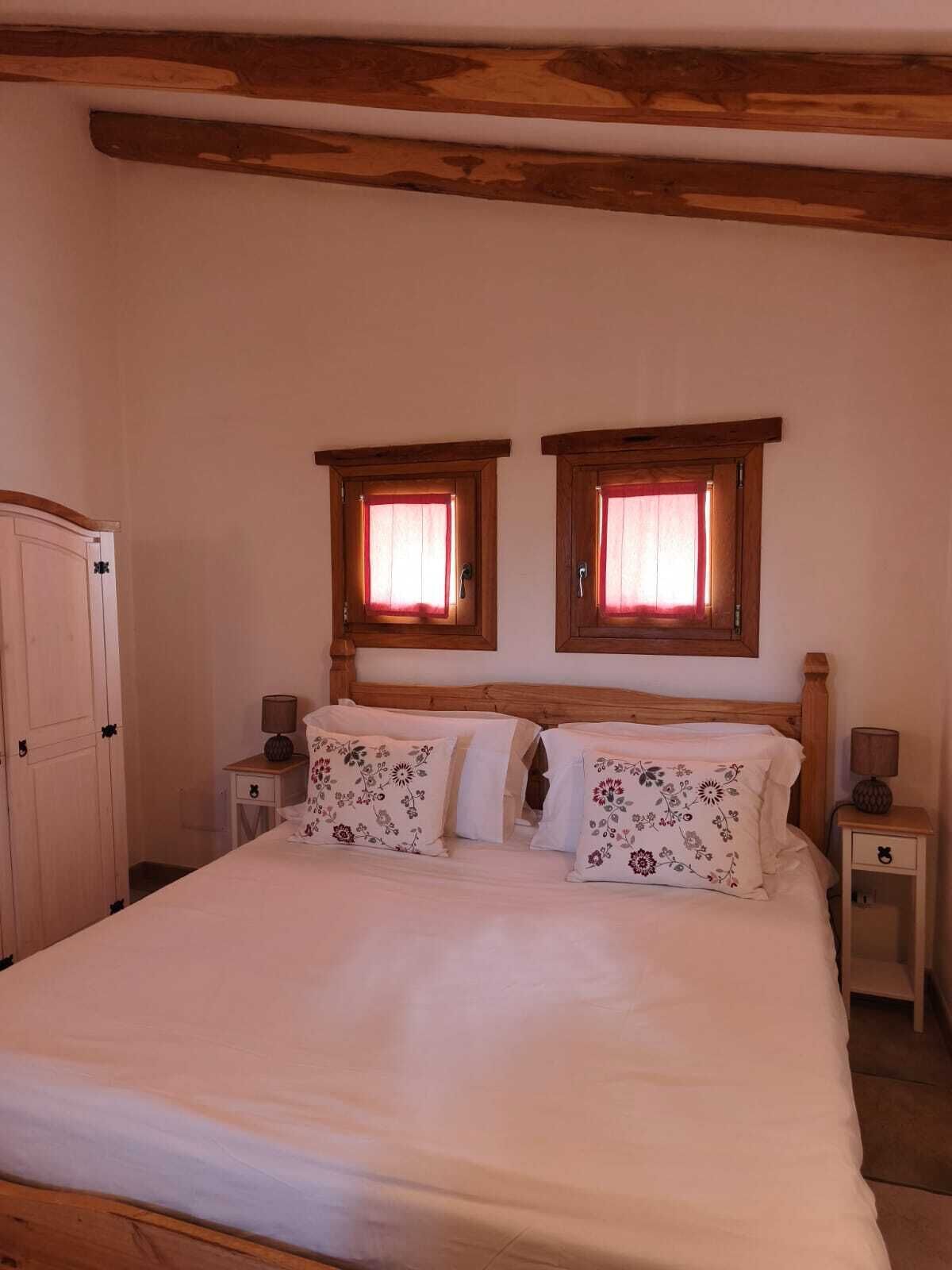 A bedroom with a wooden bed frame, two bedside tables with lamps, a wardrobe, and two windows with curtains.