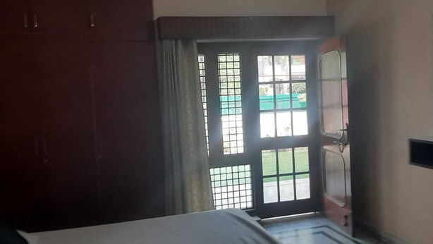 A room with a bed, a wardrobe, a window with a view of a green area, and a small shelf. - Hotel Shivdham (Lucknow)