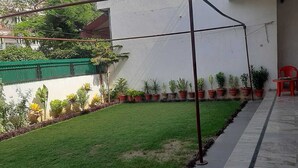 A courtyard with a lawn, potted plants, a red chair, and a white building. - Hotel Shivdham (Lucknow)