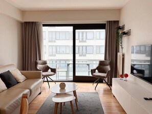 A modern living room with a sofa, two armchairs, a coffee table, a TV, and a balcony with a view of buildings. - Cozy Coastal Apartment With Dual Terraces (Middelkerke)