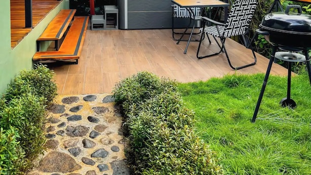 En udendørs terrasse med grill, siddepladser og en stenhusvej, der fører til indgangen.