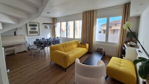 Un salon moderne avec un canapé jaune, un pouf blanc et un coin repas agrémenté d’un piano.
