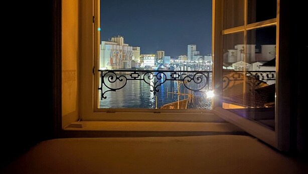 Une chambre avec vue sur le paysage urbain nocturne, un balcon avec une balustrade et une fenêtre donnant sur l’eau.