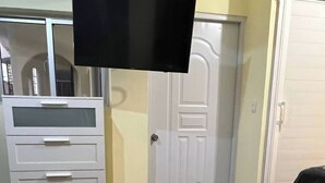 Una habitación con piso de madera, un armario blanco con cajones, una puerta cerrada y un televisor montado en la pared.
