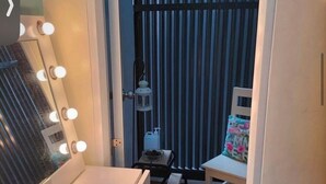 Ein Zimmer mit Schminkspiegel, Beleuchtung, einem Sessel, einem kleinen Tisch und einem Fenster mit Jalousien.
