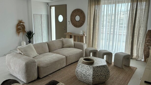 A modern living room with a beige sofa, a round coffee table, and a large window with sheer curtains.