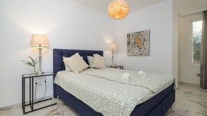 Un dormitorio con cama, dos mesitas de noche, lámpara colgante, una obra de arte en la pared y ventana.