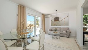 Un moderno salón con una mesa de comedor de vidrio, sofás blancos, una lámpara colgante y un amplio ventanal con cortinas.