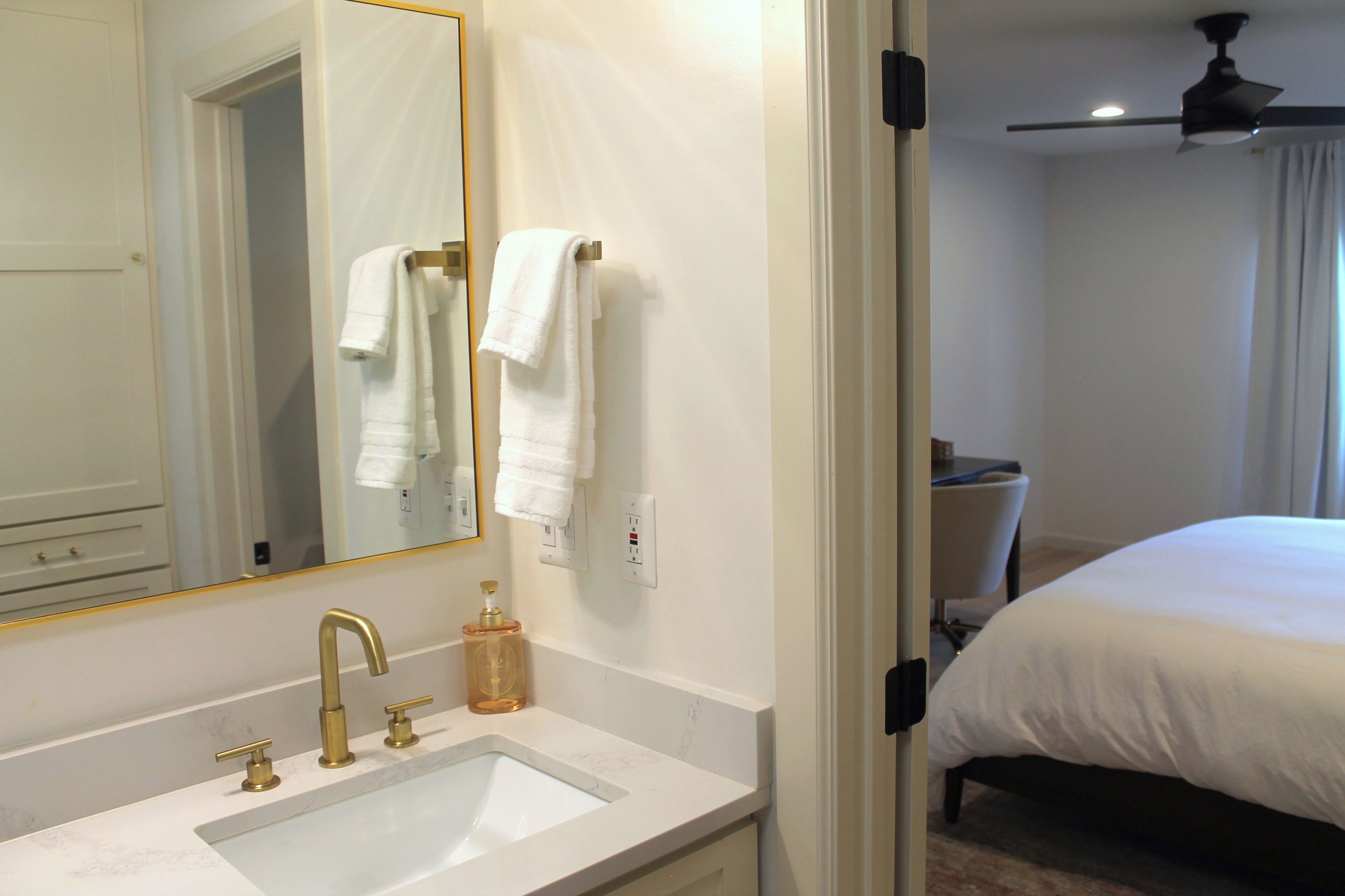 A hotel bathroom with a sink, mirror, towel rack, and a bed in the adjacent room.