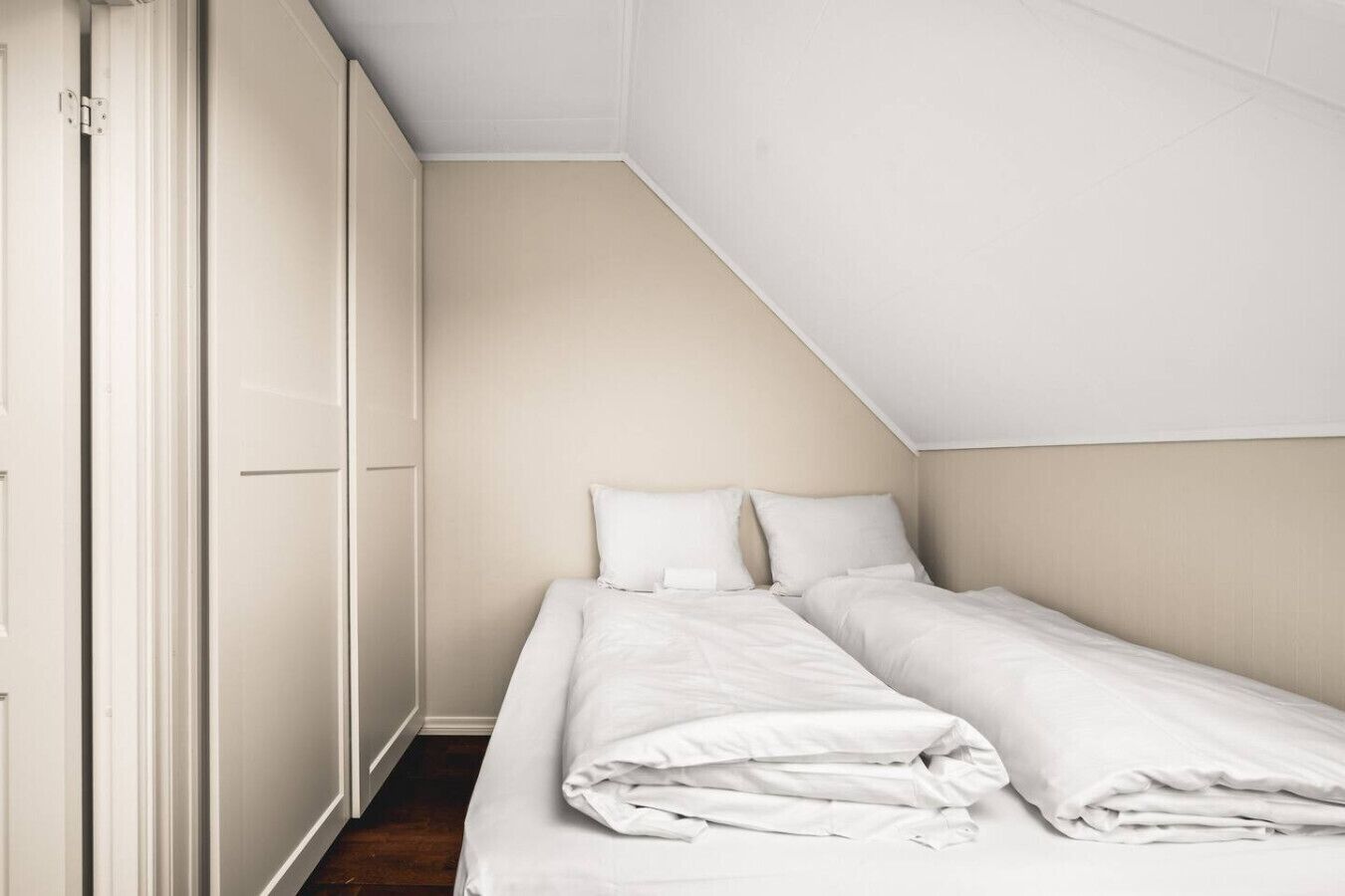 Ein kleines Zimmer mit einem Einzelbett, schrägen Wänden, weißen Wänden und einer Tür links.