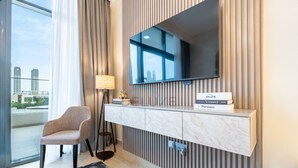 A modern hotel room with a large window, a flat-screen TV mounted on a wooden panel, a plush chair, and a console table with a nameplate. - Cozy Modern Studio Just A Quick Drive from Meydan Racecourse, Azizi Riviera 20 (Dubai)