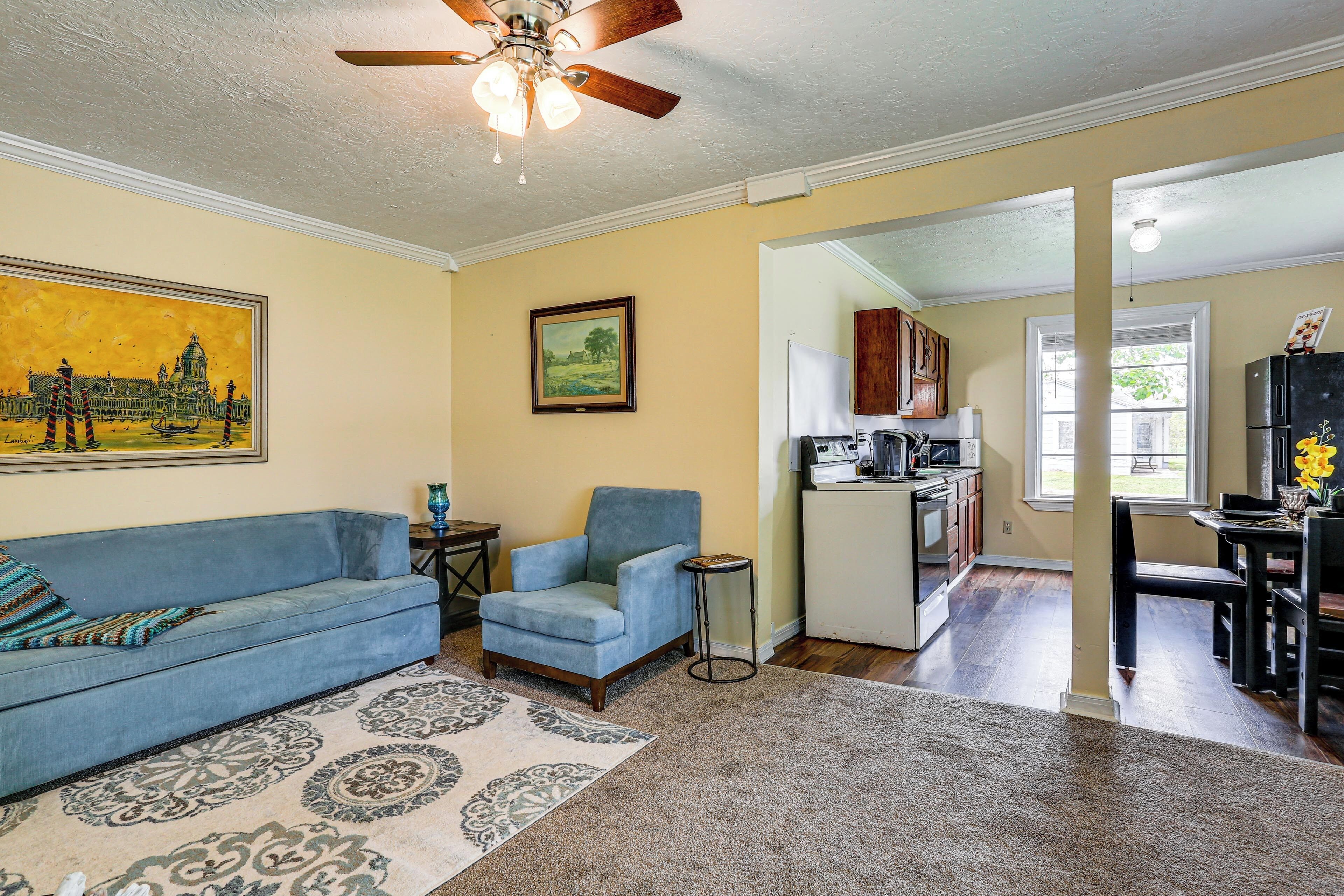 A living room with a blue sofa, a small armchair, a ceiling fan, a framed painting, and a kitchen in the background.