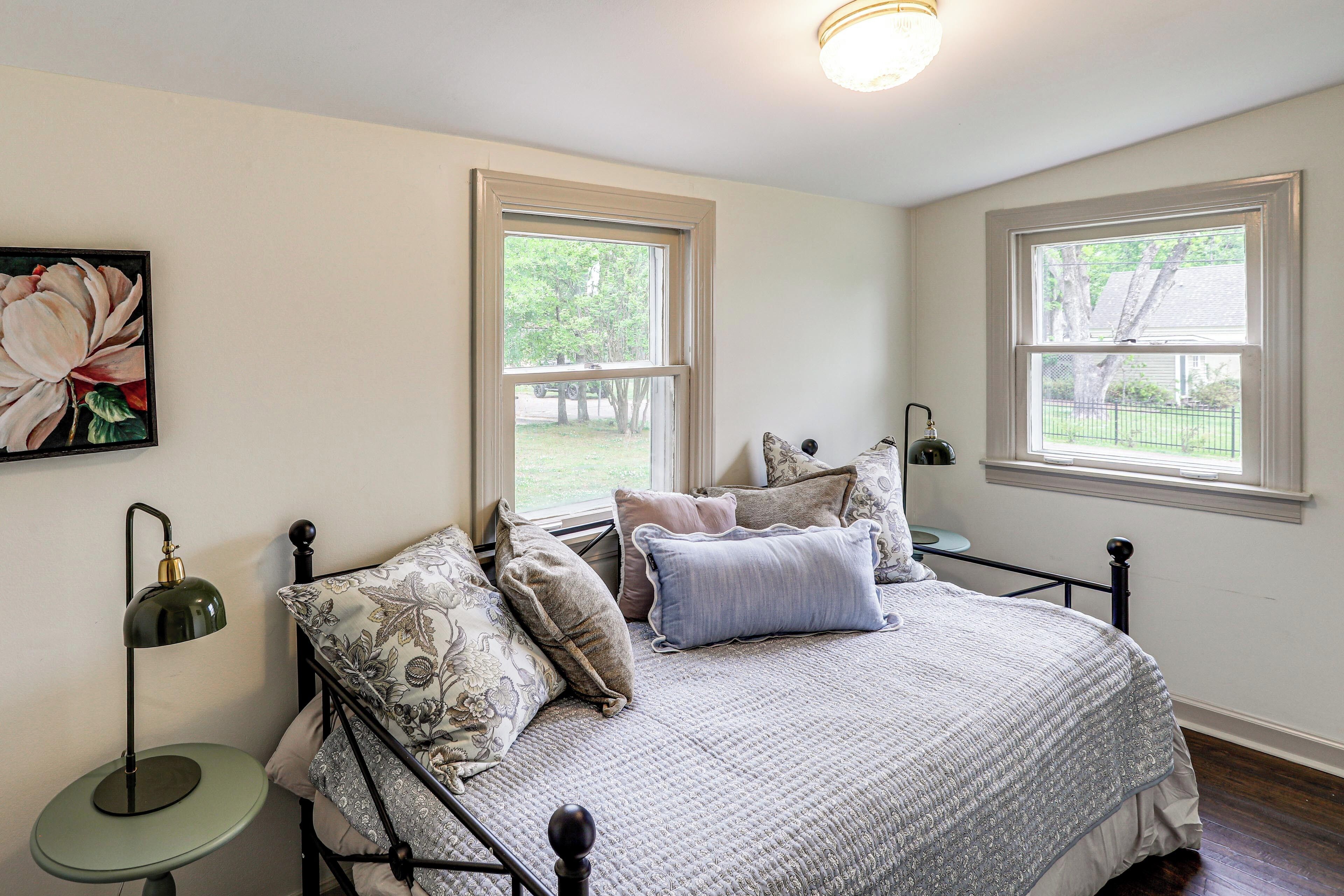 A bedroom with a bed, two windows, a nightstand, a lamp, and a floral painting.