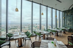 A restaurant with large windows, green chairs, and a city view.