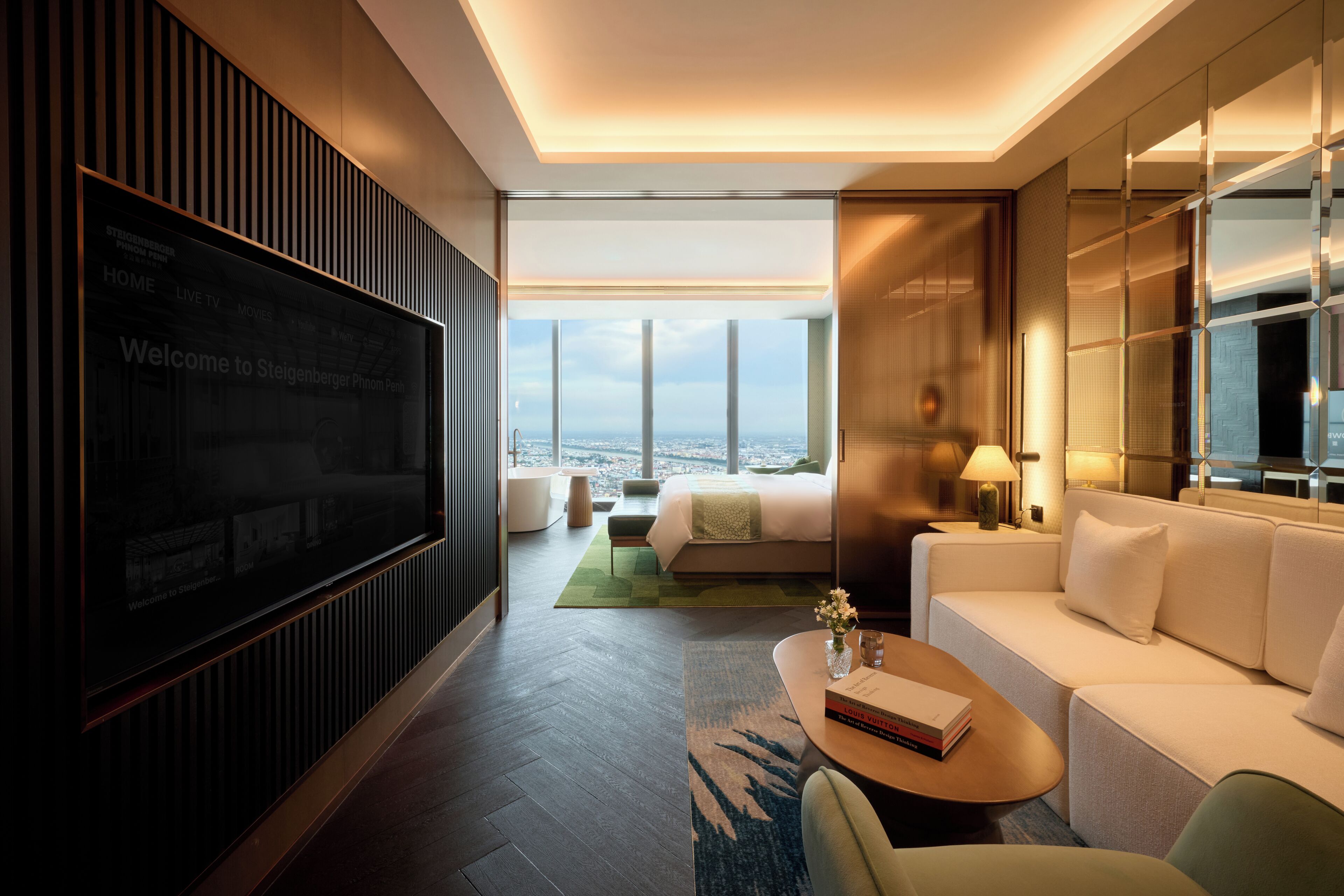 A modern hotel room with a large TV, a sofa, a coffee table, and a view of the city.