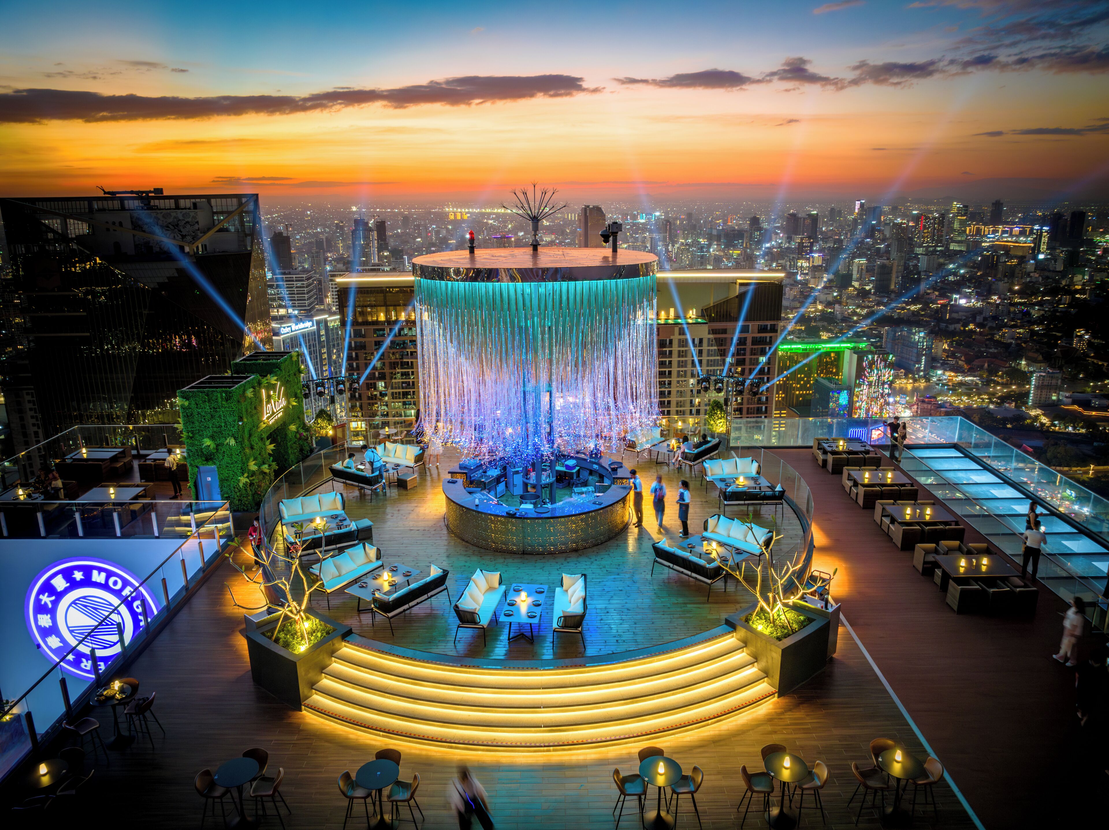 Un bar en la azotea con un área de estar circular, una fuente de agua y vistas a la ciudad al atardecer.
