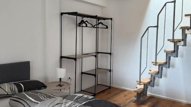 A modern bedroom with a bed, a hanging clothes rack, a bedside table with a lamp, and a wooden staircase.