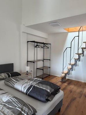 A modern bedroom with a bed, a hanging clothes rack, a bedside table with a lamp, and a wooden staircase. - Stettenblick Apartments (Loerrach)