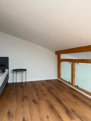 A room with a bed, a small round table, wooden flooring, and a large window with a wooden frame. - Stettenblick Apartments (Loerrach)