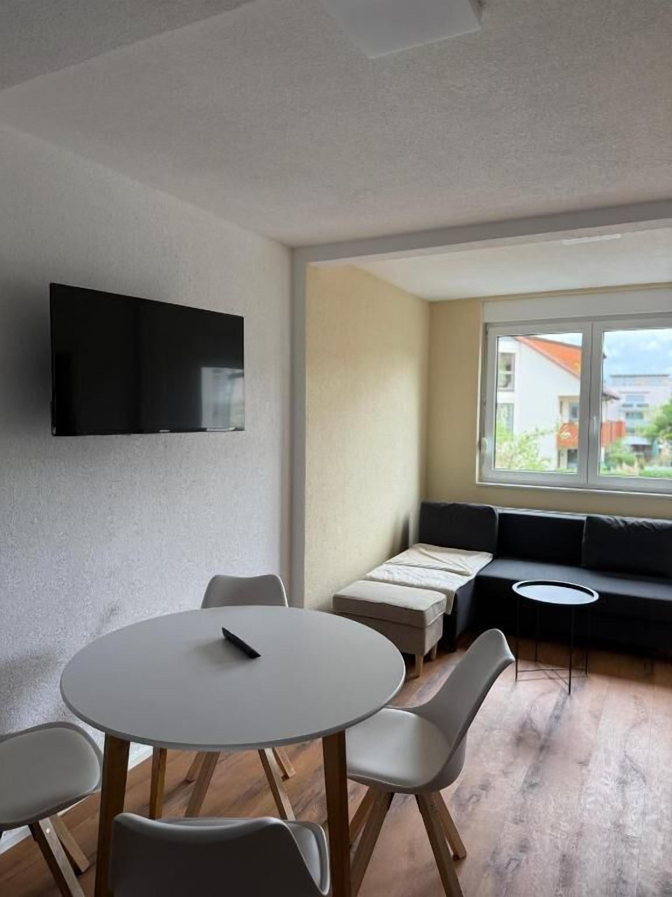 A modern living room with a round table, chairs, a sofa, a TV mounted on the wall, and a large window.