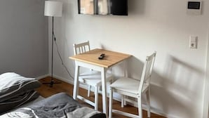 A bedroom with a bed, a bedside table, a desk with chairs, a TV mounted on the wall, and a floor lamp.