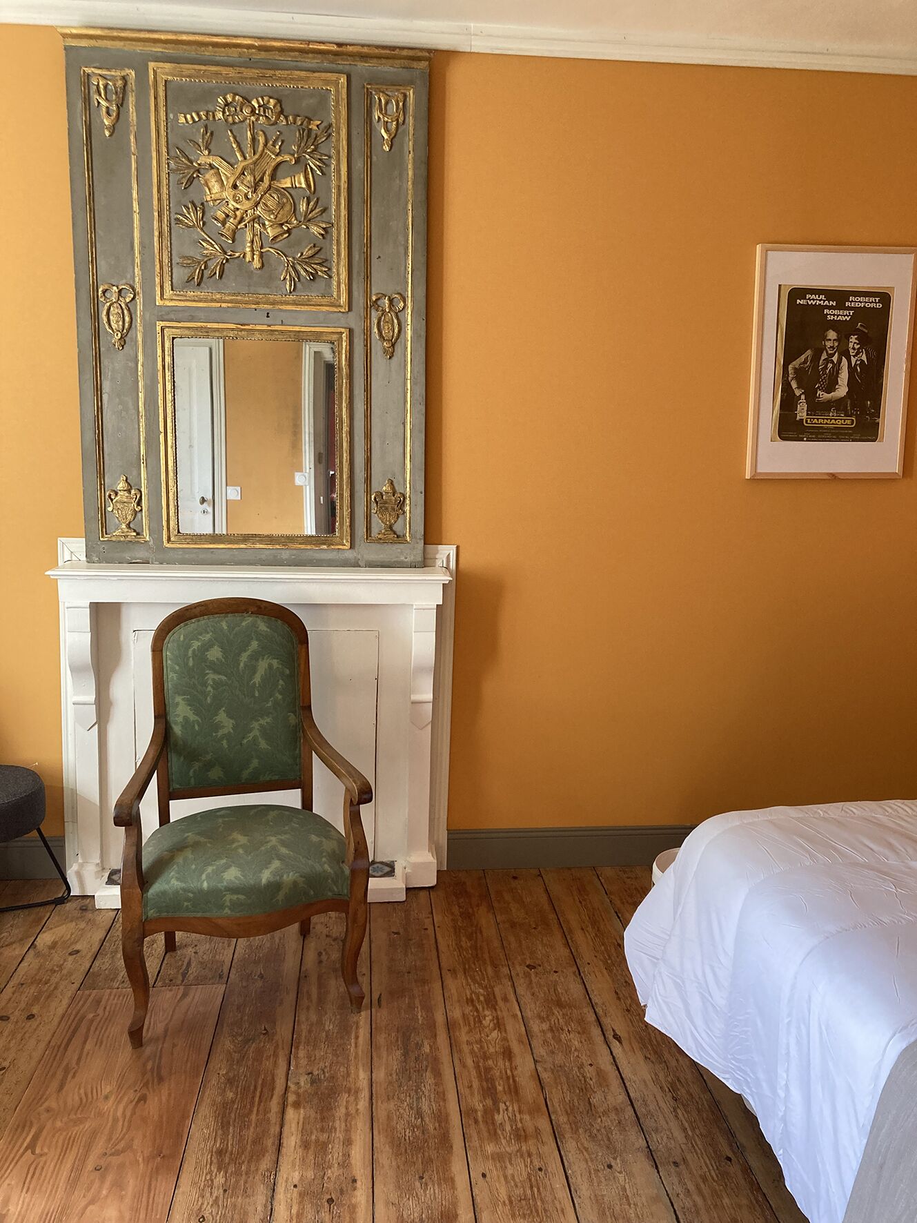 Une pièce avec un plancher en bois, un fauteuil recouvert d’un tissu vert, un lit blanc, une cheminée agrémentée d’un miroir et un tableau encadré accroché au mur.