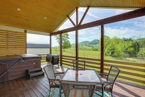A covered outdoor deck with a table and chairs, a hot tub, and a view of a green landscape.