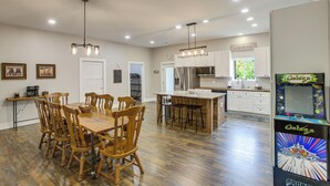 A spacious kitchen with a dining area, wooden chairs, a central island, and a retro arcade machine. - Sunset Soaks & Family Fun: Rural Tennessee Retreat (Bethpage)