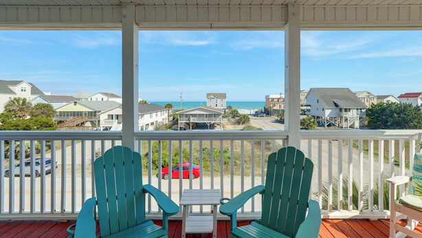 Een overdekt balkon met twee groene Adirondack-stoelen en een kleine tafel, met uitzicht op een strand en woongebied.