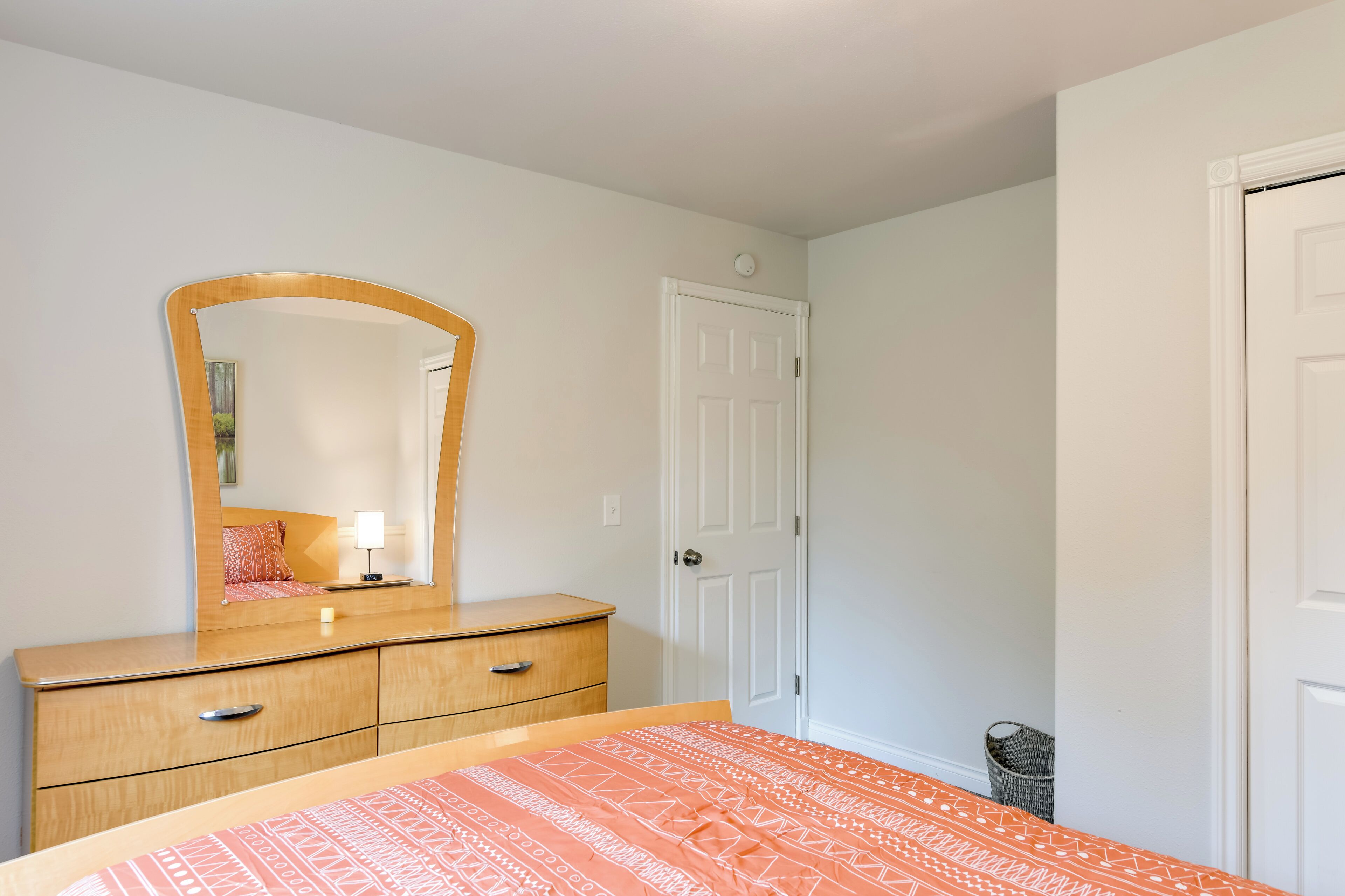 A bedroom with a wooden dresser, a mirror, a bed with an orange patterned bedspread, a white door, and a small trash bin.