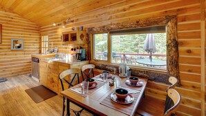 Een houten chalet van binnen, met een eettafel gedekt voor vier personen, een keukenhoek en een raam met uitzicht op een terras.