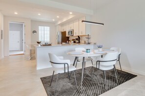 A modern kitchen with white cabinets, a central island, and a dining area with white chairs and a round table. - Golf + Poolside Fun! Extended Base in Ibis Landing (Lehigh Acres)