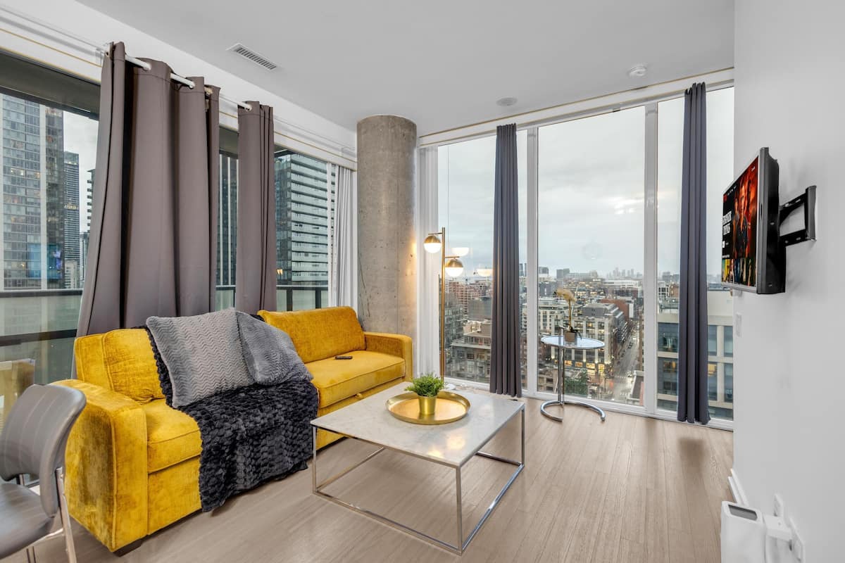 A modern living room with a yellow sofa, a coffee table, and a large window with a city view.