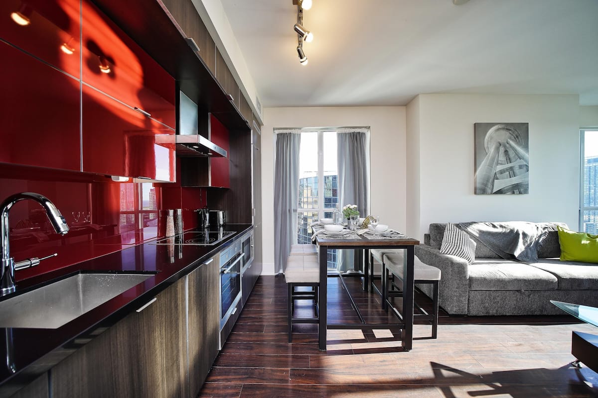 A modern kitchen with a red backsplash, a dining area with a bench, and a living room with a sofa and a painting on the wall.
