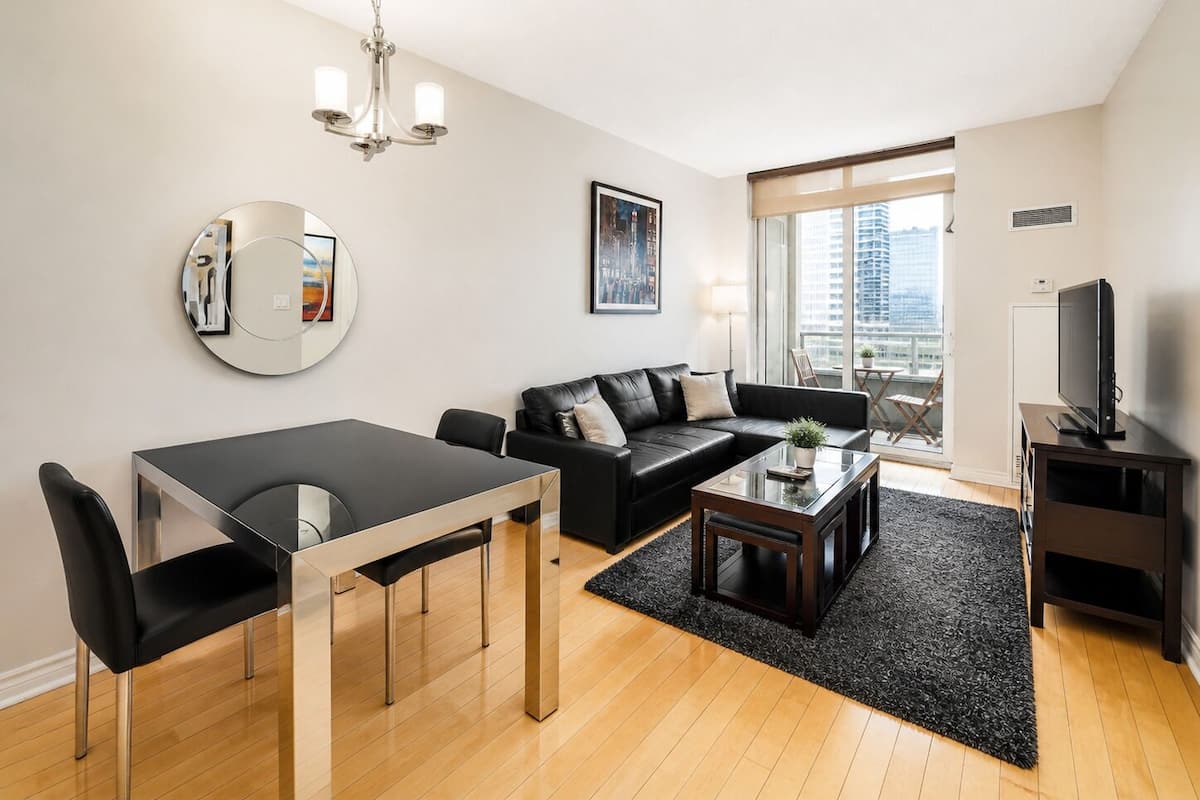 A modern living room with a dining area, a black leather sofa, a round mirror, and a television.