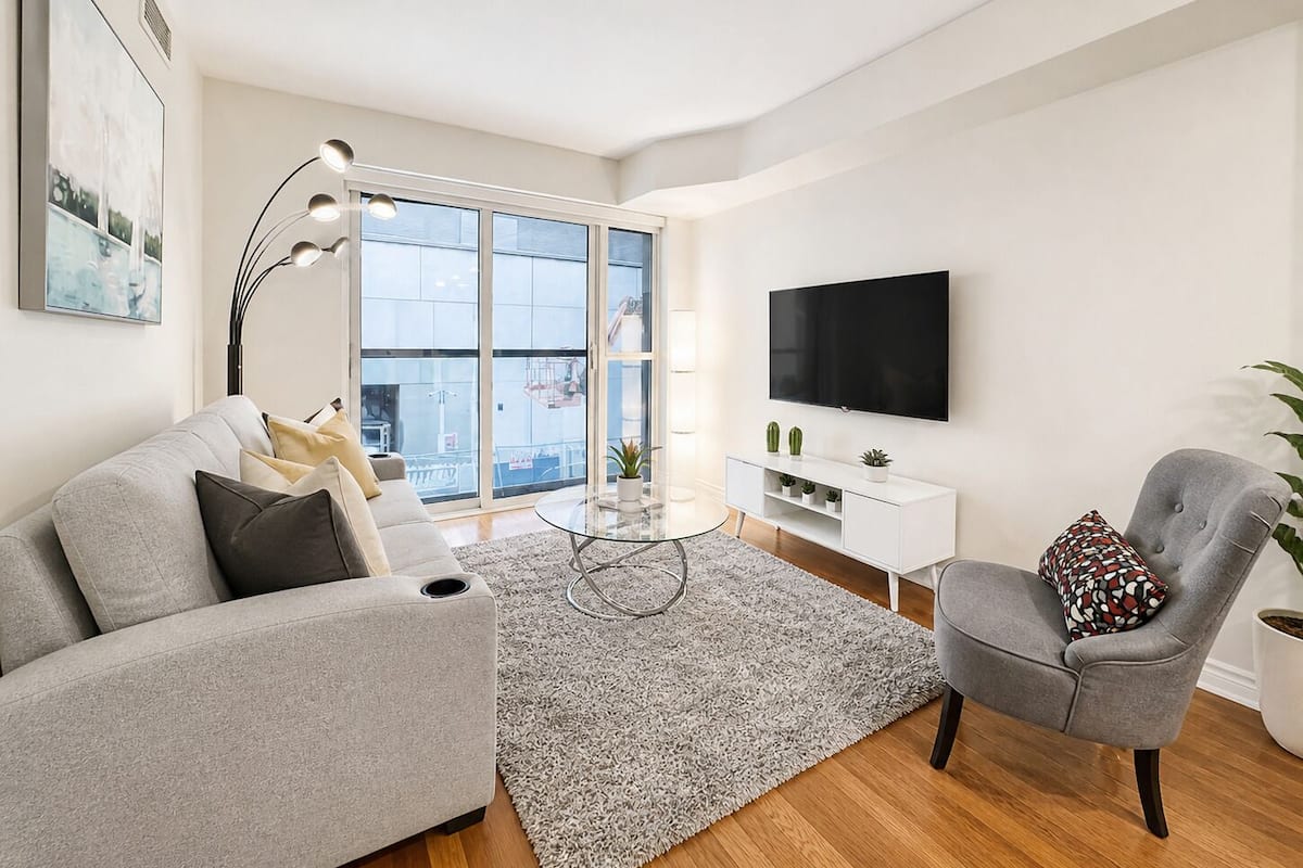 A modern living room with a grey sofa, a round coffee table, a grey armchair, a white TV stand, and a large window with a view of buildings.