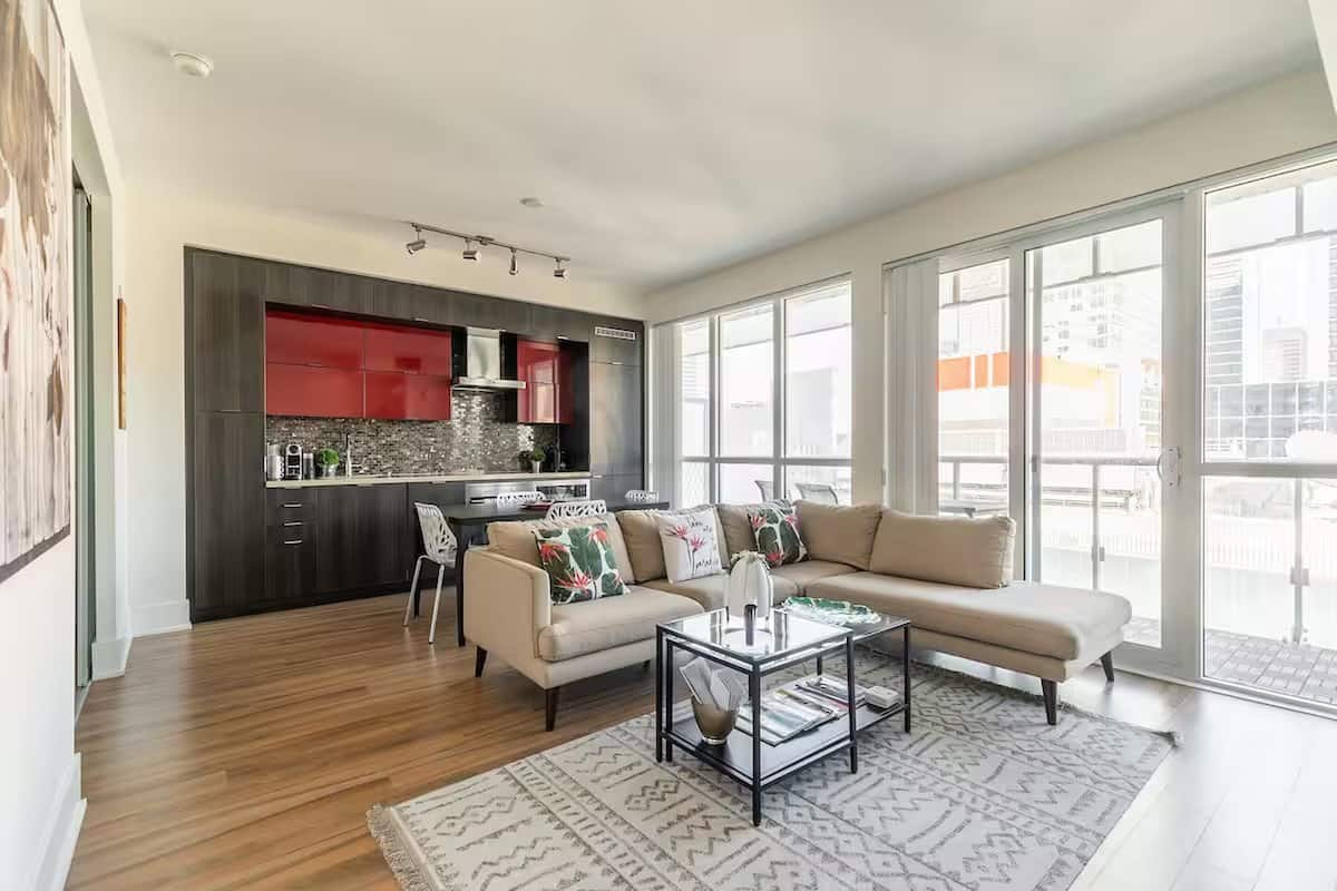 A modern living room with a sofa, coffee table, and a kitchen area with red cabinets and a window.