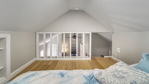 Una camera da letto con soffitto spiovente, pavimento in legno, un letto con biancheria a motivi e vista su una stanza adiacente tramite un divisorio bianco.