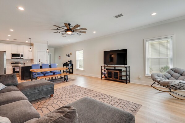 A modern living room with a sofa, ottoman, and a rocking chair. There is a kitchen area with a microwave, stove, and dining table. A ceiling fan and a TV are also visible. - NEW! Foley Family Escape w/ Fenced Yard (Foley)