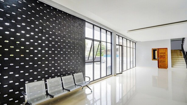 Un espace d’attente moderne avec un mur noir perforé, des chaises en métal, de grandes fenêtres et une porte en bois.