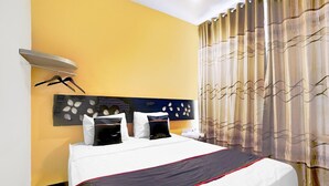 A hotel room with a bed, a headboard with a decorative panel, a wall-mounted shelf with a towel, and a curtain with a wavy pattern.