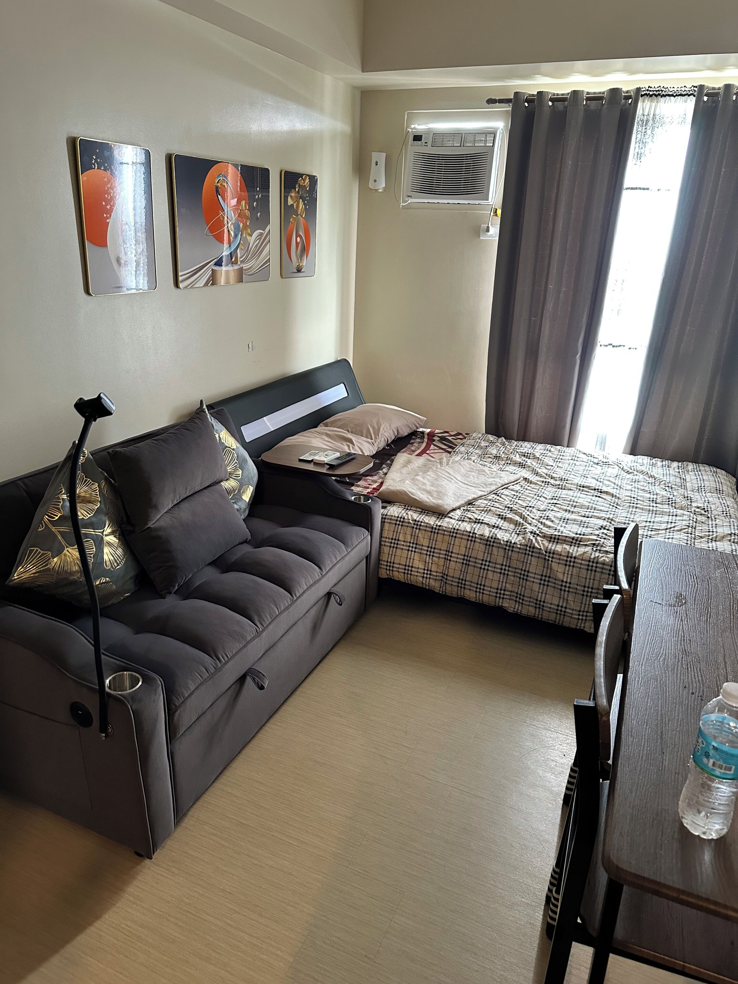 A compact hotel room with a sofa, a bed, a table, and an air conditioning unit.