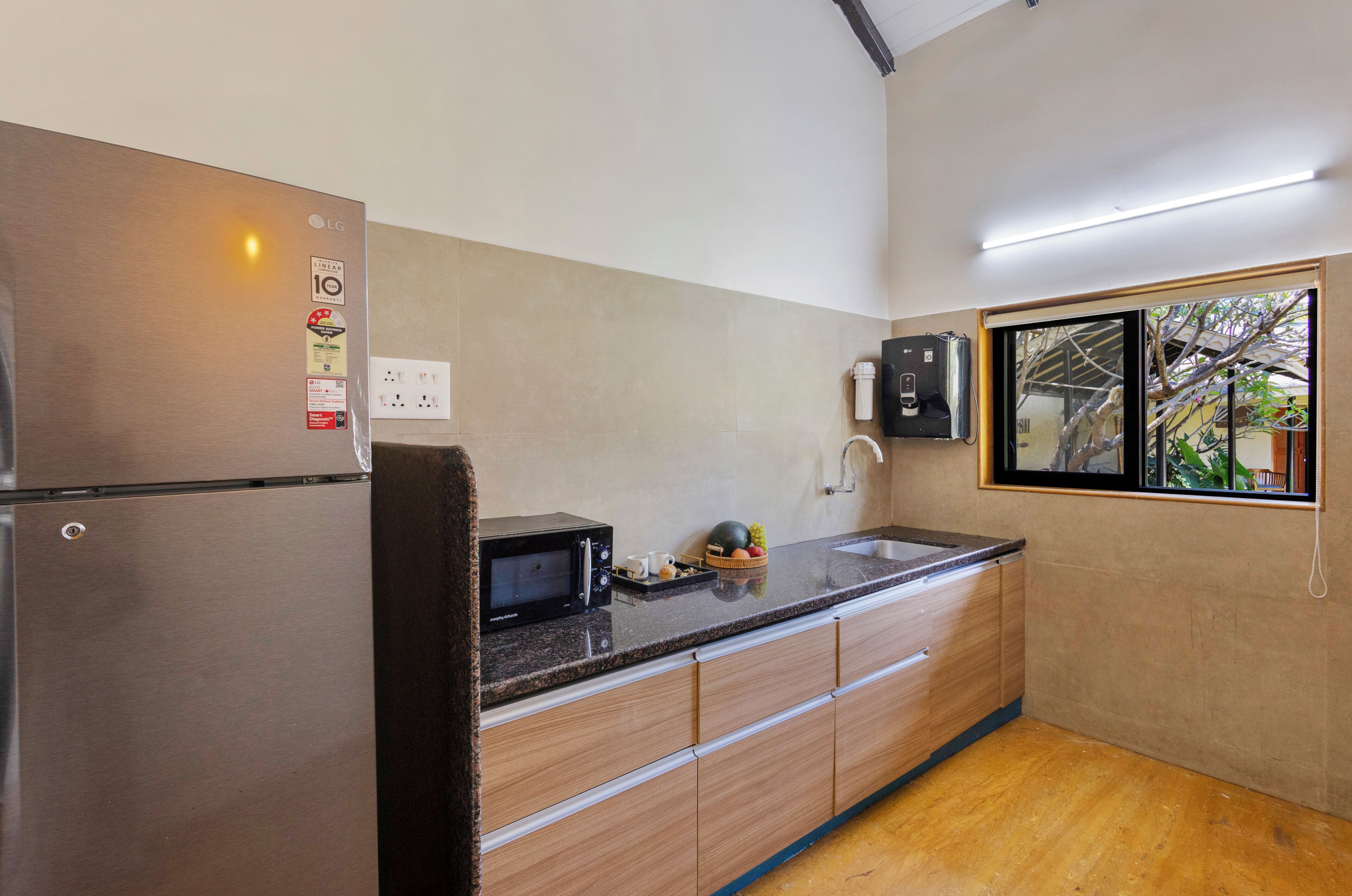 A kitchen with a stainless steel refrigerator, a microwave, wooden cabinets, a granite countertop, and a window with a view of trees.