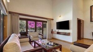 A living room with a sofa, chairs, a coffee table, a flat-screen TV mounted on the wall, and a large window with curtains.