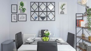 Un coin repas avec une table blanche, des chaises noires, un miroir à cadre noir, des œuvres d’art encadrées et une bibliothèque contenant des livres et des objets décoratifs.
