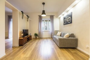 Ein Wohnzimmer mit einer Couch, einem Fernseher, Holzboden, einer hängenden Pflanze und einem Fenster mit Vorhängen.