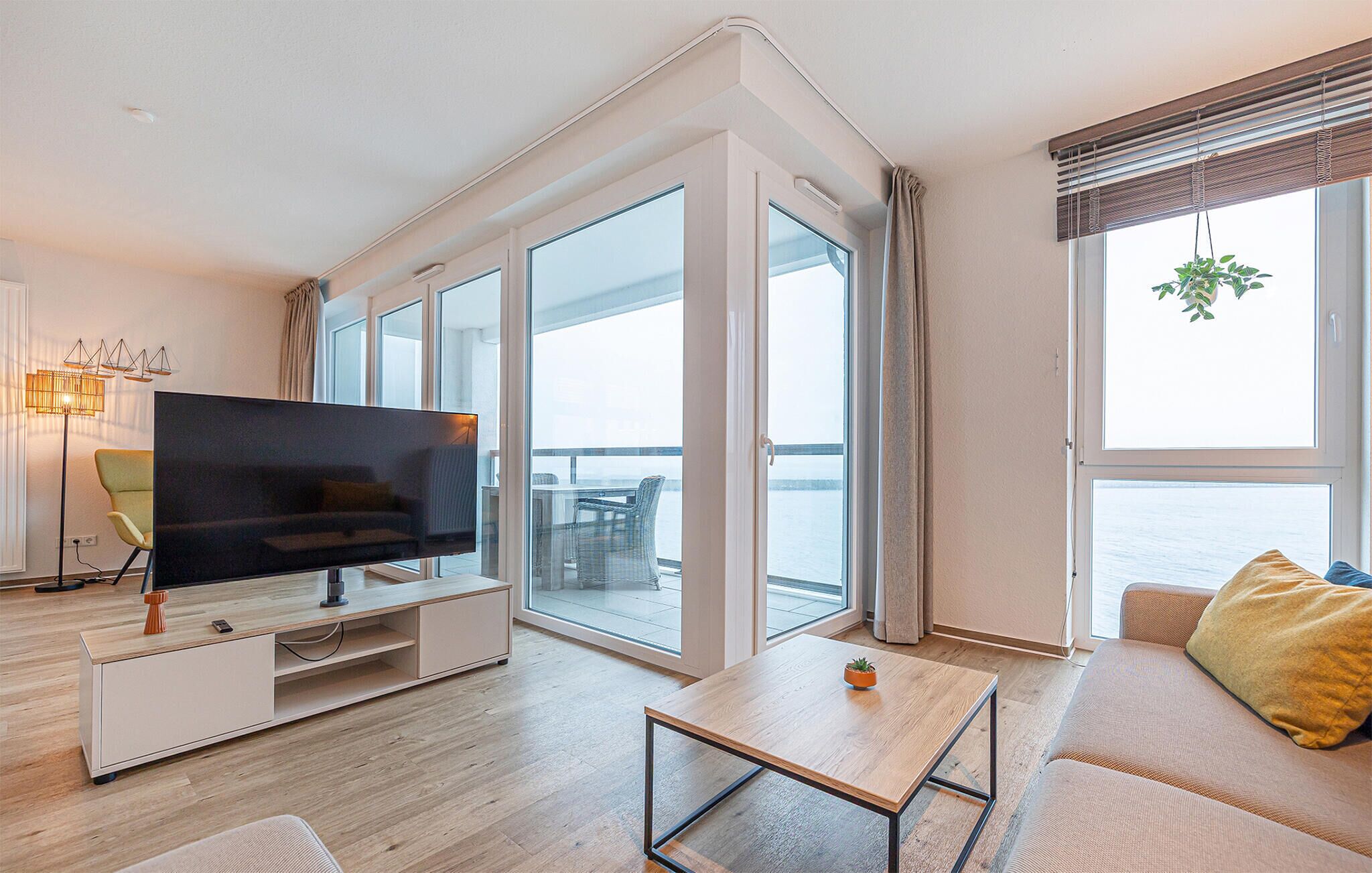 A modern living room with a flat-screen TV, a sofa, a coffee table, and large windows offering a view of the sea.