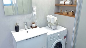 Un baño moderno con un lavamanos blanco, lavadora integrada, un espejo y repisas con toallas y artículos de aseo.